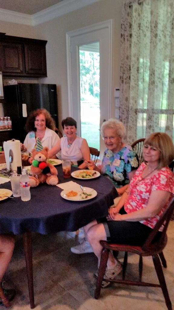 Four women smiling at the dining table with a stuffed teddy bear seated as a fun table guest