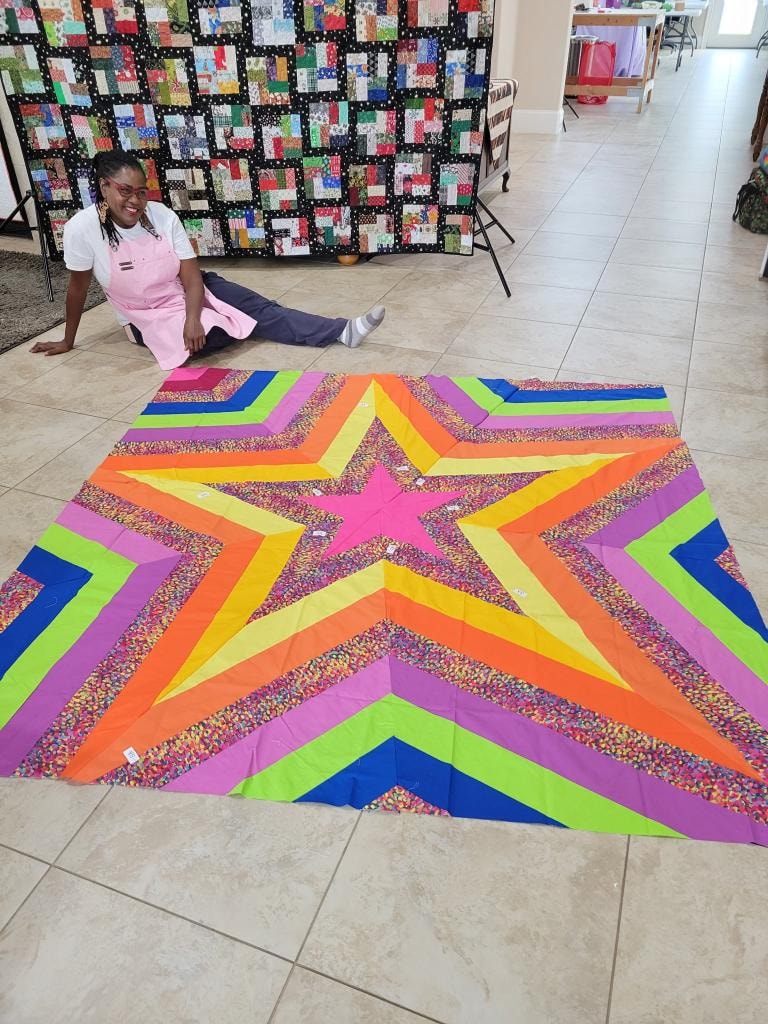 Guest posing beside a large vibrant star quilt in pink, orange, yellow, lime green, blue, and purple laid out on the floor
